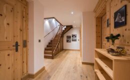 Interior view of a modern wooden hallway featuring a staircase, wooden doors, and decorative wall art. The warm-toned wooden finishes and soft lighting create a cozy atmosphere.