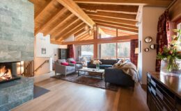Cozy living room with high wooden ceilings, large windows, and a stone fireplace, featuring comfortable sofas, a coffee table, and decorative flowers, perfect for a warm and inviting atmosphere.