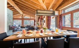 Cozy dining area with a wooden ceiling, featuring a beautifully set table with a variety of breakfast foods and drinks, surrounded by large windows that provide natural light and scenic views.
