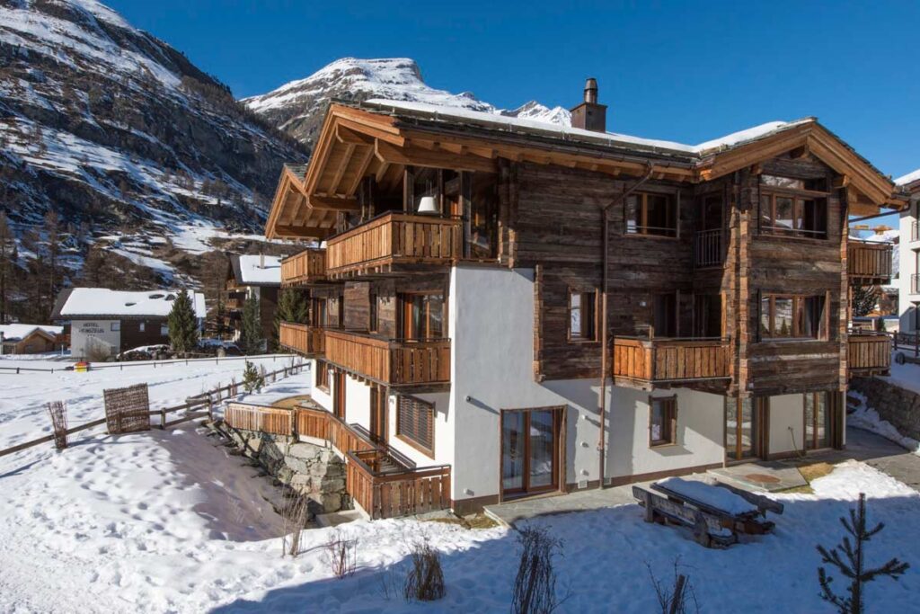 Charming wooden chalet in a snowy landscape, surrounded by mountains, featuring multiple balconies and large windows for natural light. Ideal for winter getaways and outdoor activities. | Only on SopranoVillas