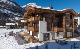 Charming wooden chalet in a snowy landscape, surrounded by mountains, featuring multiple balconies and large windows for natural light. Ideal for winter getaways and outdoor activities.