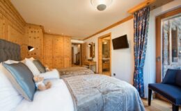 Cozy hotel room featuring two beds with plush bedding, wooden paneling, and a stylish chair, illuminated by natural light from a window with colorful curtains.