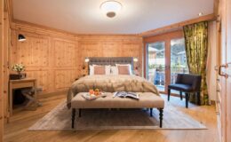 Luxurious hotel room featuring a king-sized bed with plush bedding, wooden paneling, a cozy seating area, and large windows with green curtains, creating a warm and inviting atmosphere.