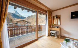 Cozy hotel room with wooden decor, featuring large windows that offer a stunning view of the Matterhorn mountain and a private balcony. A small desk and modern amenities are visible, creating a welcoming atmosphere for guests.