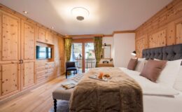 Cozy hotel bedroom featuring wooden paneling, a plush bed with decorative pillows, a stylish ottoman, and a balcony view. The room includes modern amenities like a flat-screen TV and elegant lighting, creating a warm and inviting atmosphere.