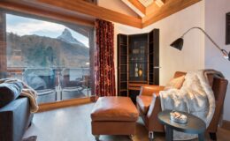 Cozy living room interior featuring a leather armchair, wooden flooring, and large windows with mountain views. Warm lighting and decorative elements create a inviting atmosphere.