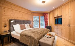 Cozy modern bedroom featuring wooden walls, a plush bed with decorative pillows, a stylish ottoman, and a TV unit, illuminated by warm lighting and large windows.