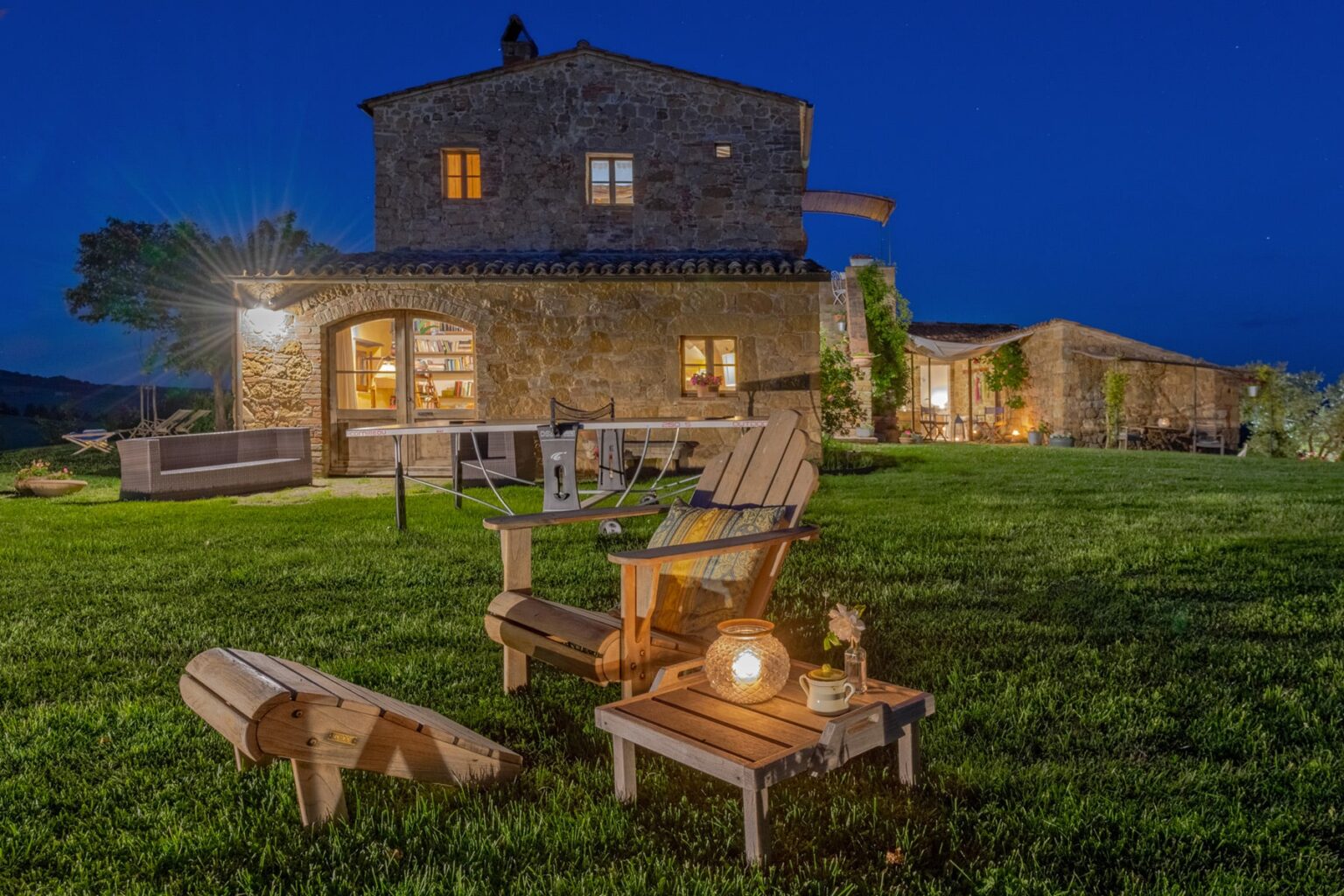 Cozy stone house at dusk with illuminated windows, surrounded by lush green grass. Outdoor seating area features wooden chairs and a lantern, creating a serene atmosphere for relaxation. Ideal for vacation rentals or countryside retreats. | Only on SopranoVillas