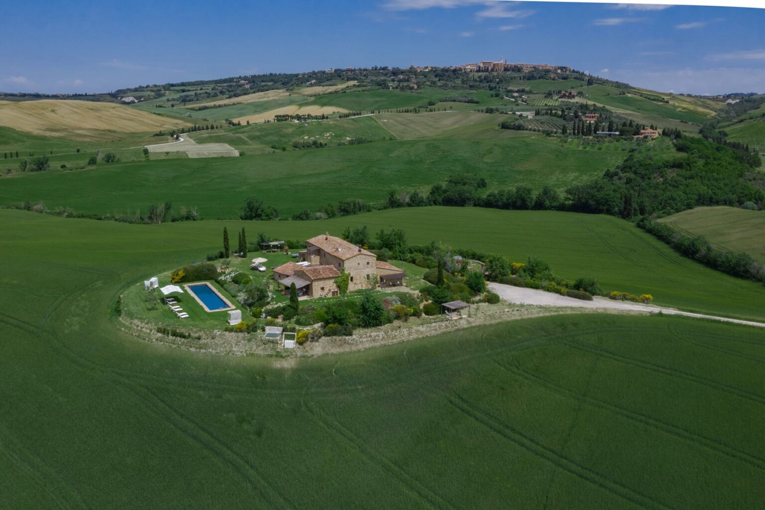Aerial view of a picturesque countryside villa surrounded by lush green fields, featuring a swimming pool, landscaped gardens, and a winding driveway. The scenic landscape includes rolling hills and a clear blue sky, highlighting the tranquility of rural living. | Only on SopranoVillas