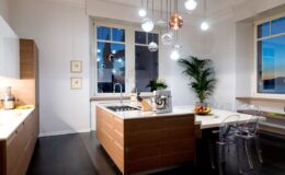 Modern kitchen interior featuring wooden cabinetry, an island with a sink, contemporary pendant lighting, and large windows with a view. The space includes clear chairs and decorative plants, creating a bright and inviting atmosphere.