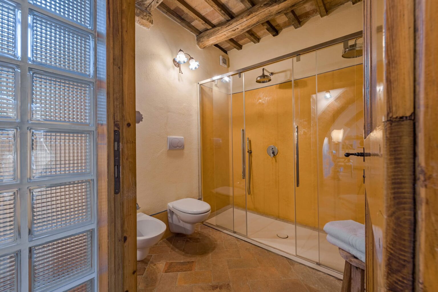 Modern bathroom interior featuring a glass shower, two toilets, and wooden beams, complemented by a warm yellow wall. Ideal for showcasing contemporary bathroom design. | Only on SopranoVillas
