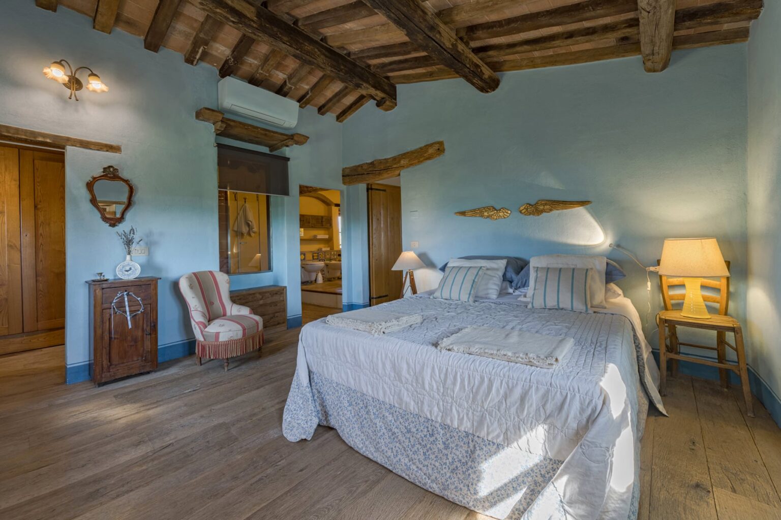 Cozy bedroom interior featuring a large bed with white linens, wooden beams on the ceiling, and warm lighting. The room includes a vintage armchair, a wooden dresser, and decorative wall mirrors, creating a serene and inviting atmosphere. | Only on SopranoVillas