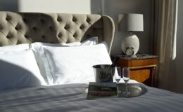 Luxurious hotel bedroom featuring a tufted headboard, crisp white linens, a silver tray with champagne flutes and a bucket, a bedside lamp, and a wooden nightstand. Natural light enhances the elegant decor.