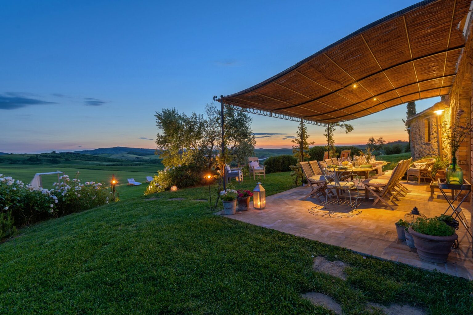 Cozy outdoor dining area with a large table and chairs under a thatched roof, surrounded by lush greenery and soft lighting, set against a scenic sunset landscape. Ideal for al fresco gatherings and enjoying nature. | Only on SopranoVillas