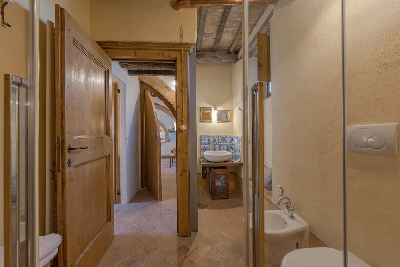 Interior view of a stylish bathroom featuring wooden doors, a glass shower enclosure, and a decorative sink area with tile accents. The warm color scheme and exposed wooden beams create a cozy atmosphere. | Only on SopranoVillas