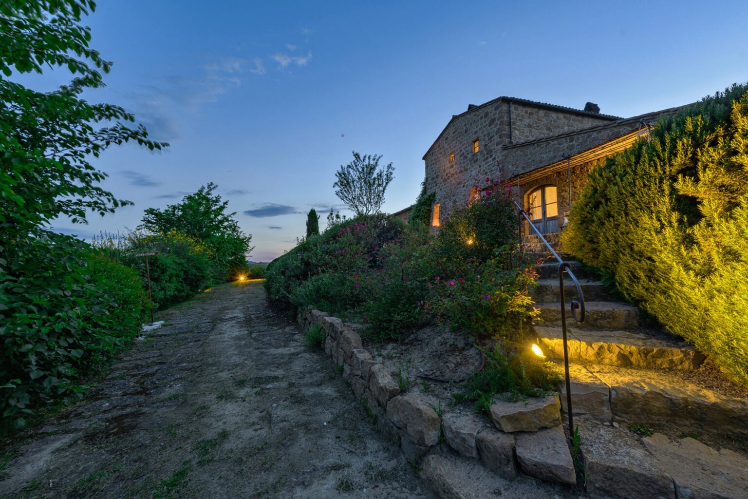 Stone house at dusk with landscaped garden and pathway illuminated by soft lighting, surrounded by greenery. | Only on SopranoVillas