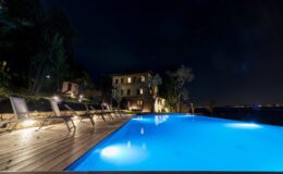 Nighttime view of a luxurious infinity pool with illuminated water, surrounded by wooden decking and lounge chairs, set against a backdrop of a scenic villa and dark sky.