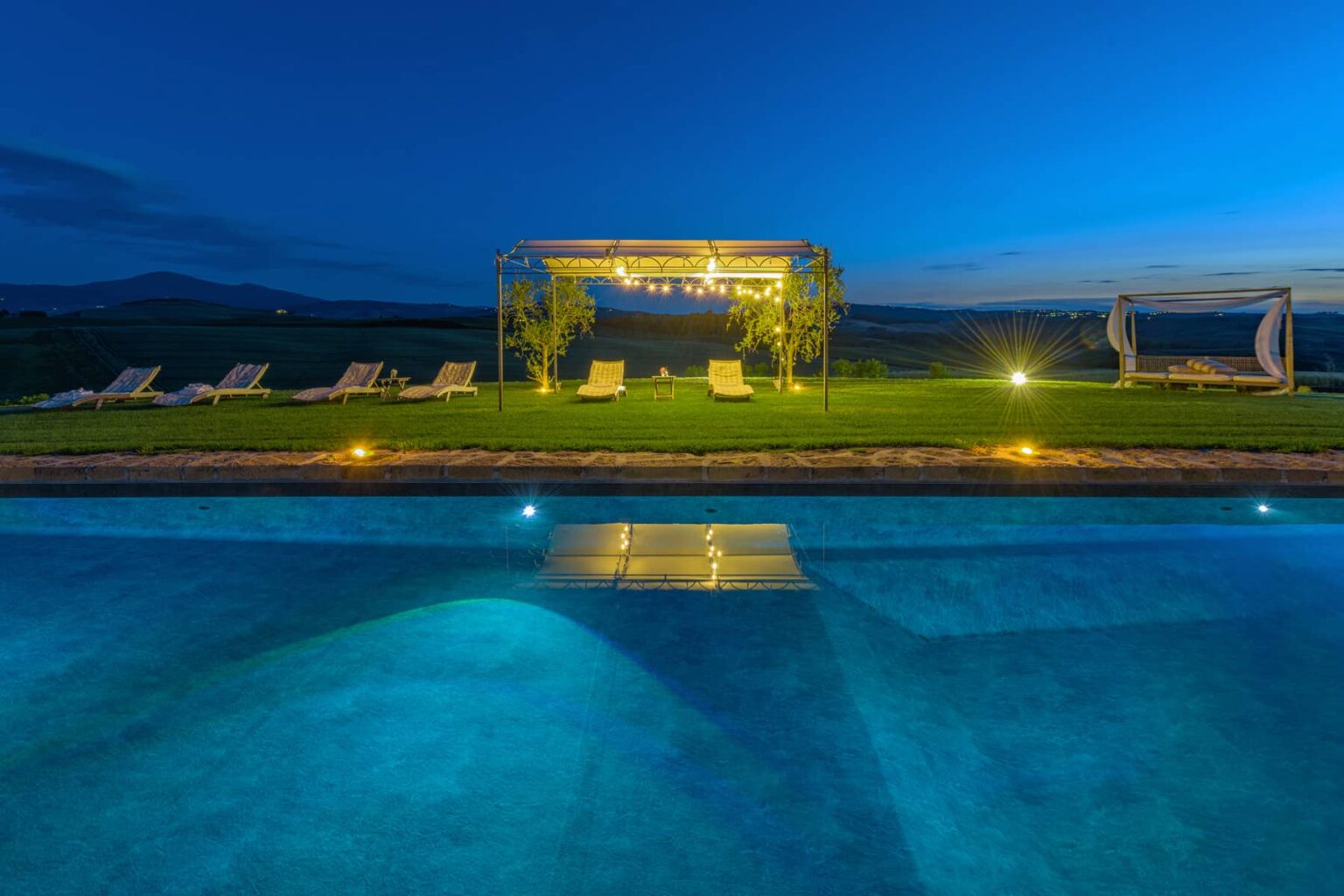 Luxury swimming pool with illuminated water at dusk, surrounded by sun loungers and a gazebo, offering a serene outdoor relaxation space. | Only on SopranoVillas