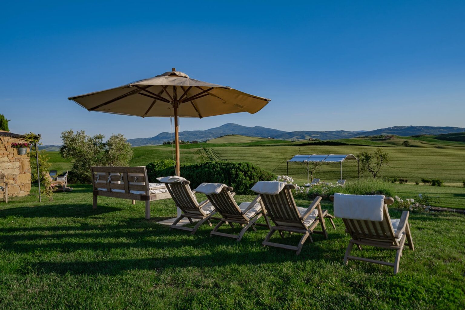 Relaxing outdoor lounge area with wooden deck chairs and a large umbrella, overlooking lush green fields and rolling hills under a clear blue sky. Ideal for enjoying nature and tranquility. | Only on SopranoVillas
