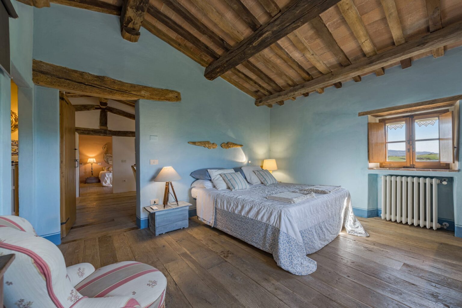 Cozy bedroom featuring a comfortable bed with soft linens, wooden beams on the ceiling, and warm lighting. The room includes a stylish chair, a radiator, and a window with picturesque views, creating a serene and inviting atmosphere. | Only on SopranoVillas