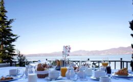 Breakfast table set with various dishes, drinks, and pastries, overlooking a serene lake and mountains under a clear blue sky. Ideal for a relaxing morning meal with a stunning view.