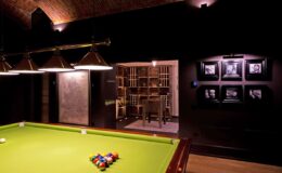 Interior view of a stylish game room featuring a green billiards table, modern lighting, and a wall adorned with framed photographs. The background includes a cozy seating area with a bookshelf, creating an inviting atmosphere for entertainment and relaxation.