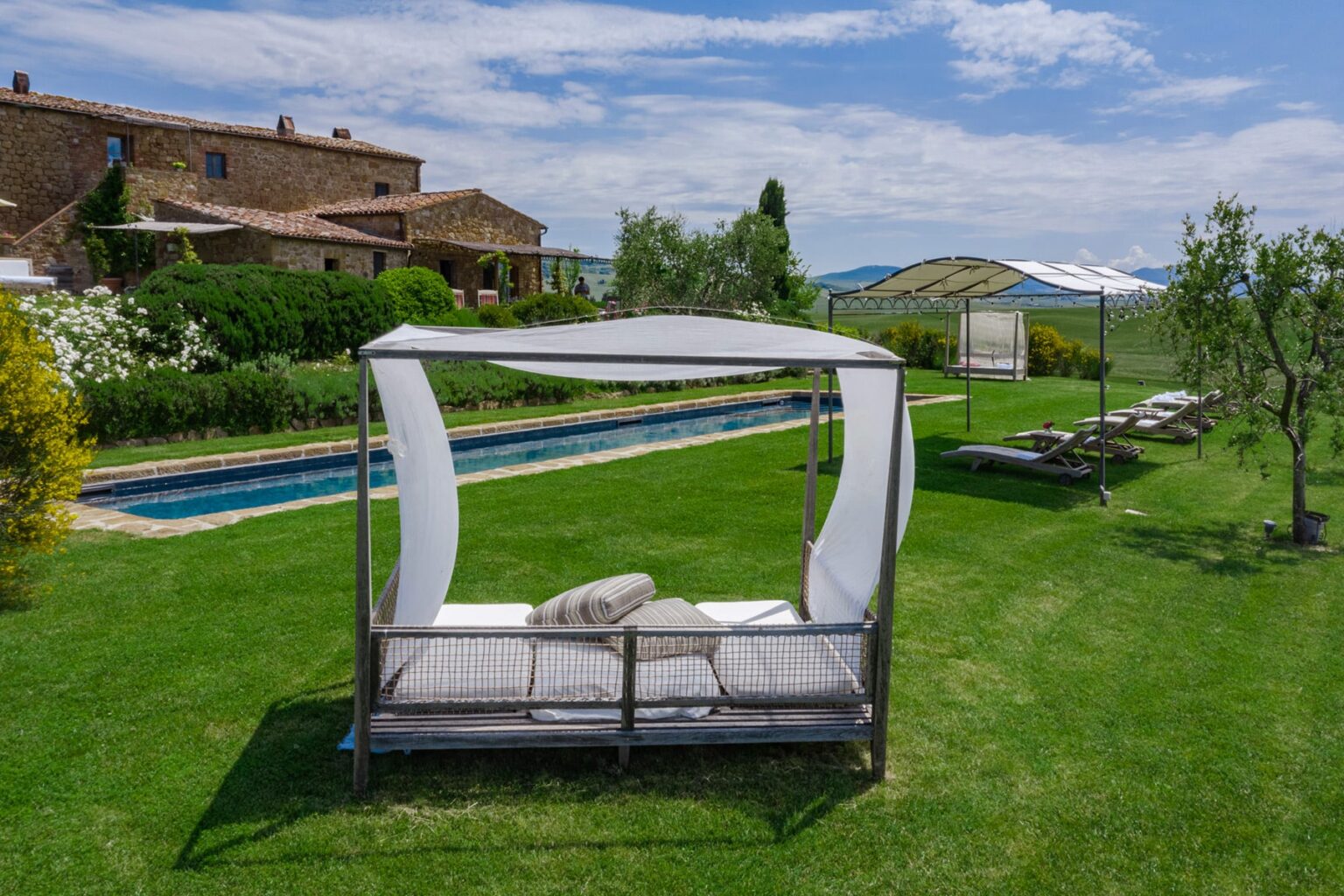 Luxurious outdoor lounge area featuring a stylish canopy bed on a lush green lawn, with a swimming pool and scenic countryside in the background. Ideal for relaxation and leisure in a serene setting. | Only on SopranoVillas
