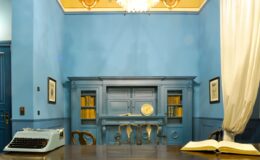 Vintage office interior featuring a blue color scheme, a classic typewriter on a wooden desk, and bookshelves filled with books. A large open book rests on the desk, while elegant drapes frame a window, adding to the room's historic charm.