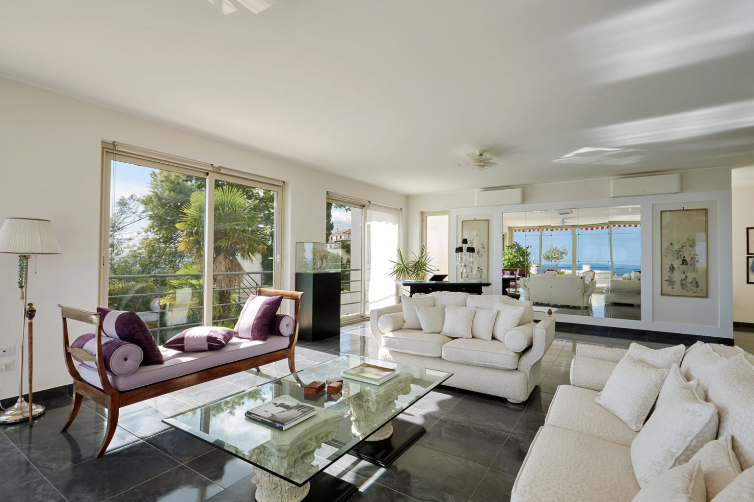 Modern living room with large windows offering a view of greenery and water, featuring a sleek glass coffee table, plush white sofas, and elegant decor elements. Bright, airy atmosphere with natural light streaming in. | Only on SopranoVillas