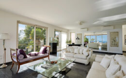 Modern living room with large windows offering a view of greenery and water, featuring a sleek glass coffee table, plush white sofas, and elegant decor elements. Bright, airy atmosphere with natural light streaming in.
