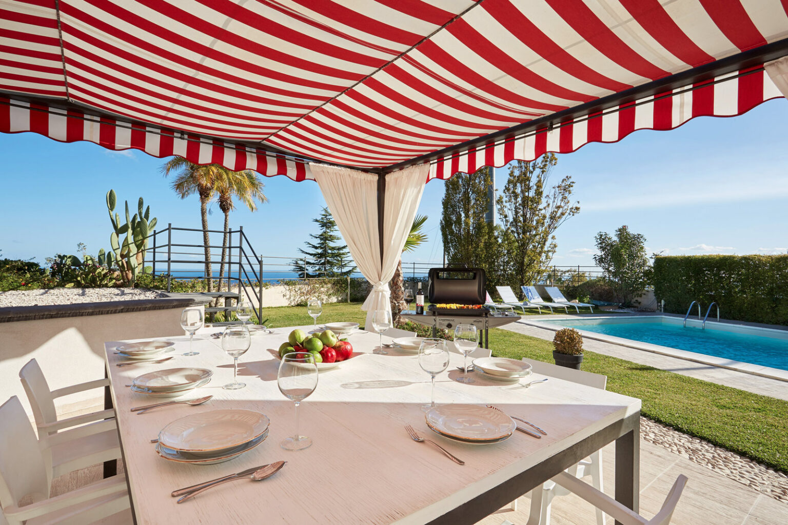 Outdoor dining area with a striped red and white awning, featuring a table set with plates and a centerpiece of fresh fruit, overlooking a swimming pool and lush greenery. Ideal for alfresco meals and gatherings in a sunny, scenic location. | Only on SopranoVillas