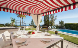 Outdoor dining area with a striped red and white awning, featuring a table set with plates and a centerpiece of fresh fruit, overlooking a swimming pool and lush greenery. Ideal for alfresco meals and gatherings in a sunny, scenic location.