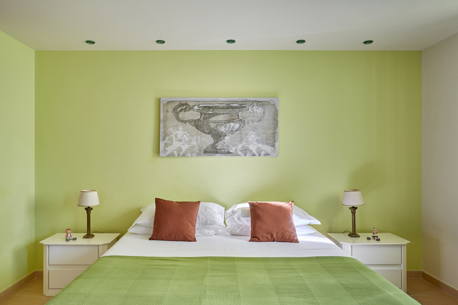 Modern bedroom featuring a green accent wall, a comfortable bed with white and orange pillows, and minimalist bedside tables with lamps. A piece of artwork hangs above the bed, enhancing the contemporary decor. | Only on SopranoVillas