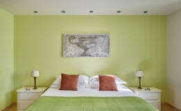 Modern bedroom featuring a green accent wall, a comfortable bed with white and orange pillows, and minimalist bedside tables with lamps. A piece of artwork hangs above the bed, enhancing the contemporary decor.