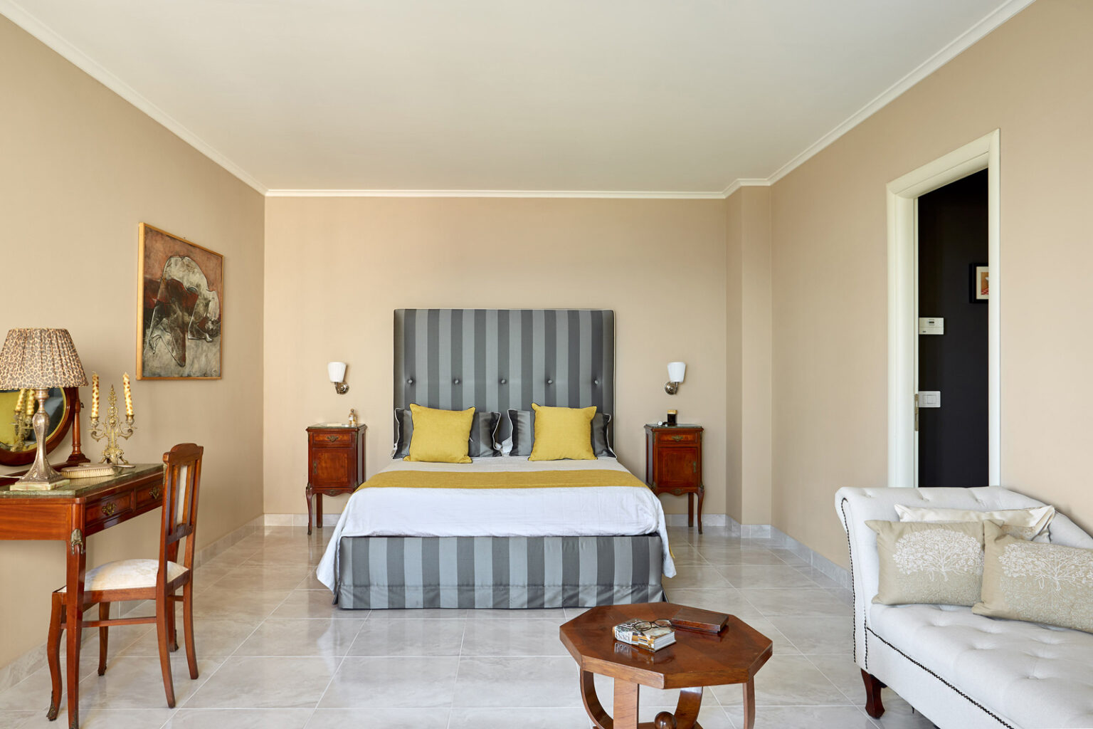 Spacious hotel room featuring a king-size bed with striped headboard, two accent pillows in yellow, elegant bedside lamps, a wooden side table, a cozy armchair, and a decorative painting on the wall, all set against a neutral beige backdrop. | Only on SopranoVillas