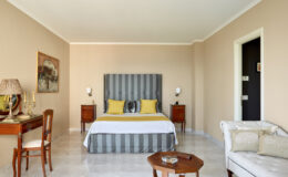 Spacious hotel room featuring a king-size bed with striped headboard, two accent pillows in yellow, elegant bedside lamps, a wooden side table, a cozy armchair, and a decorative painting on the wall, all set against a neutral beige backdrop.