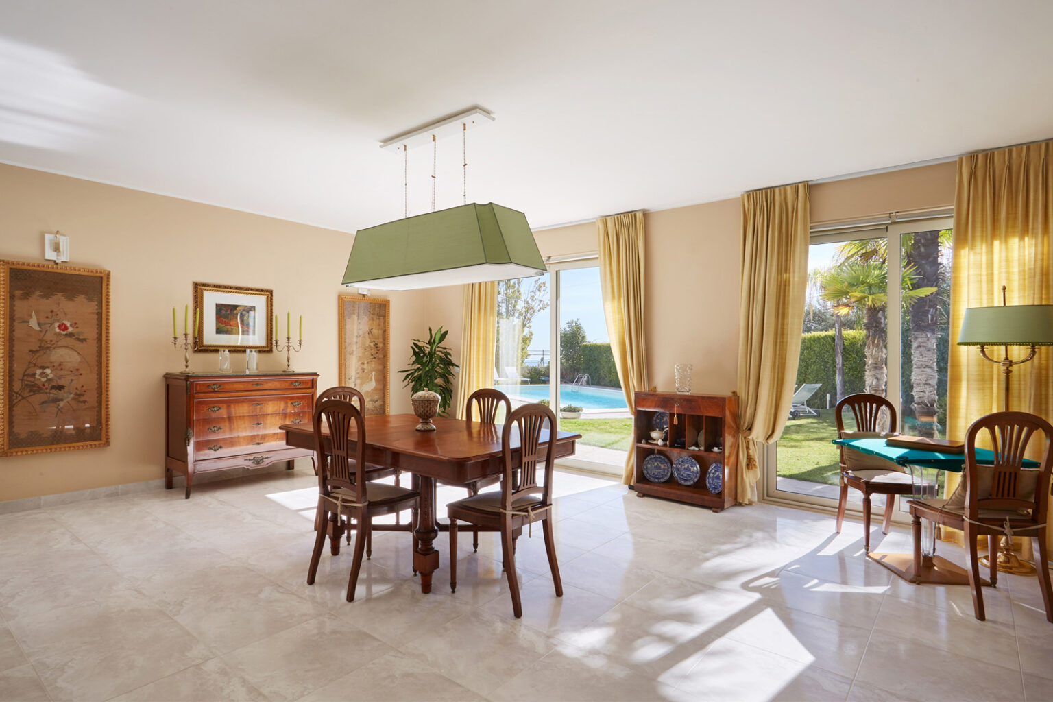 Bright and spacious dining area featuring a wooden table and chairs, elegant light fixture, and large windows with yellow curtains overlooking a pool and garden. The warm color palette and tasteful decor create a welcoming atmosphere. | Only on SopranoVillas