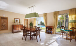 Bright and spacious dining area featuring a wooden table and chairs, elegant light fixture, and large windows with yellow curtains overlooking a pool and garden. The warm color palette and tasteful decor create a welcoming atmosphere.