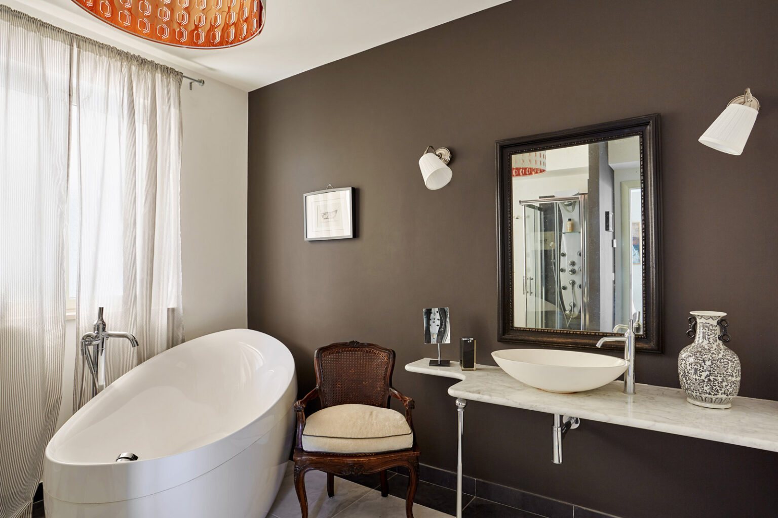 Modern bathroom featuring a freestanding bathtub, elegant marble sink, and stylish decor against a dark brown wall. Natural light filters through sheer curtains, enhancing the contemporary design. | Only on SopranoVillas