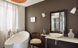 Modern bathroom featuring a freestanding bathtub, elegant marble sink, and stylish decor against a dark brown wall. Natural light filters through sheer curtains, enhancing the contemporary design.