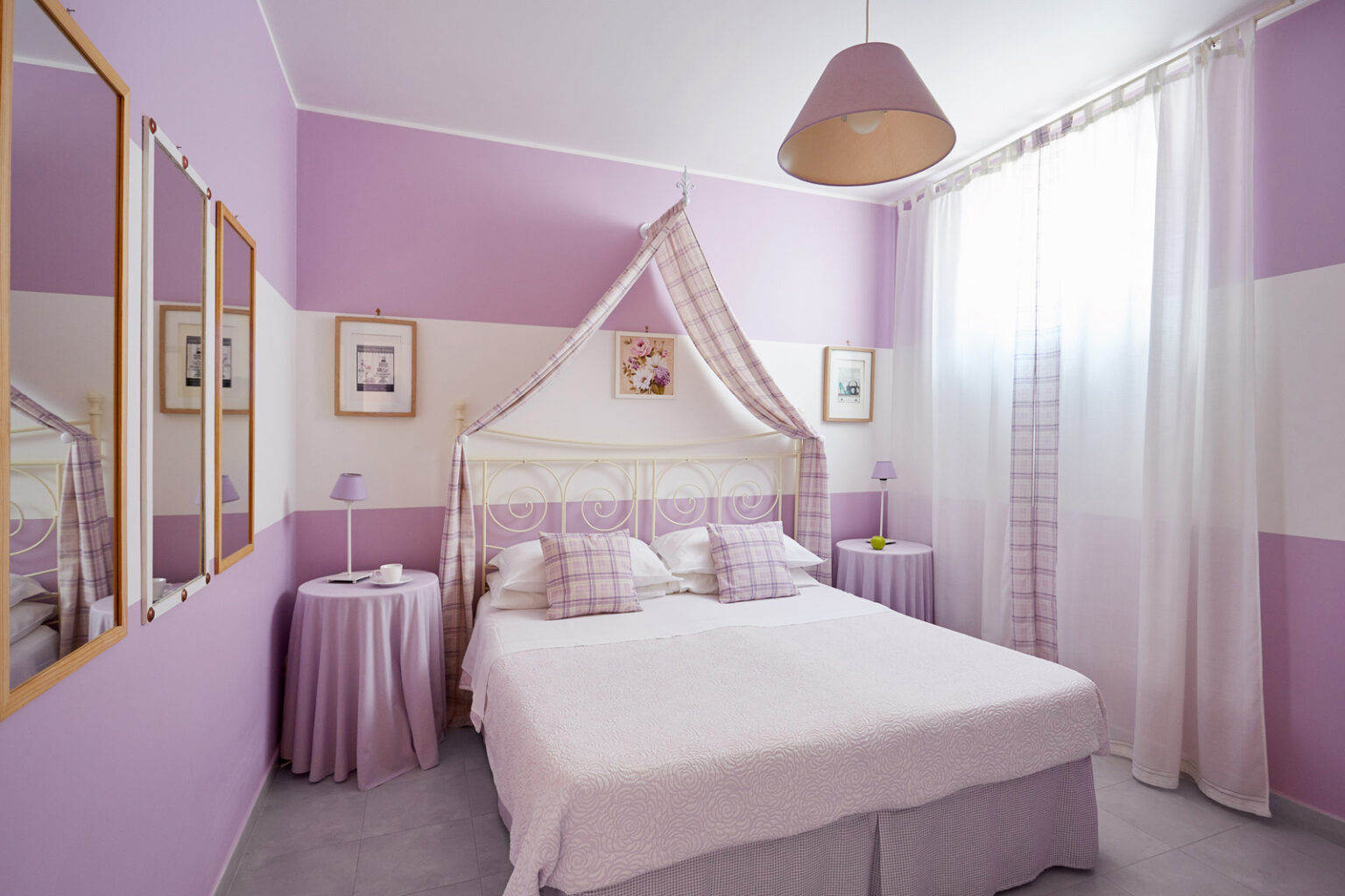 Cozy bedroom featuring a double bed with soft purple bedding, decorated walls in shades of lavender, and elegant curtains letting in natural light. The room includes stylish mirrors and a bedside table, creating a serene and inviting atmosphere. | Only on SopranoVillas