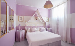 Cozy bedroom featuring a double bed with soft purple bedding, decorated walls in shades of lavender, and elegant curtains letting in natural light. The room includes stylish mirrors and a bedside table, creating a serene and inviting atmosphere.