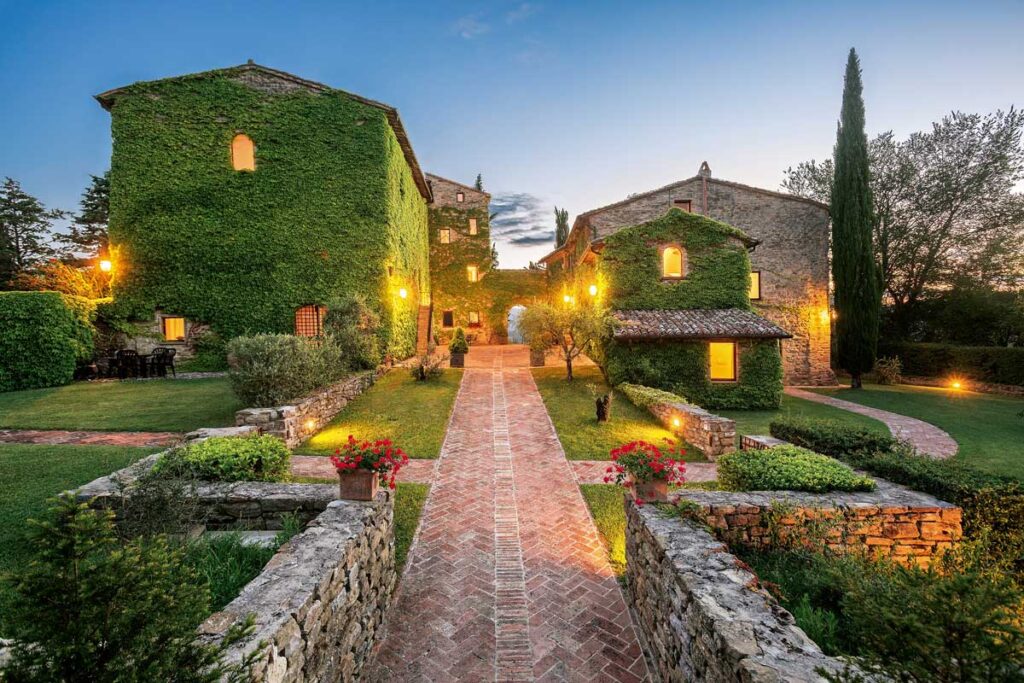Charming ivy-covered buildings illuminated at dusk, surrounded by lush gardens and a brick pathway leading to a serene outdoor setting. Ideal for romantic getaways or peaceful retreats. | Only on SopranoVillas