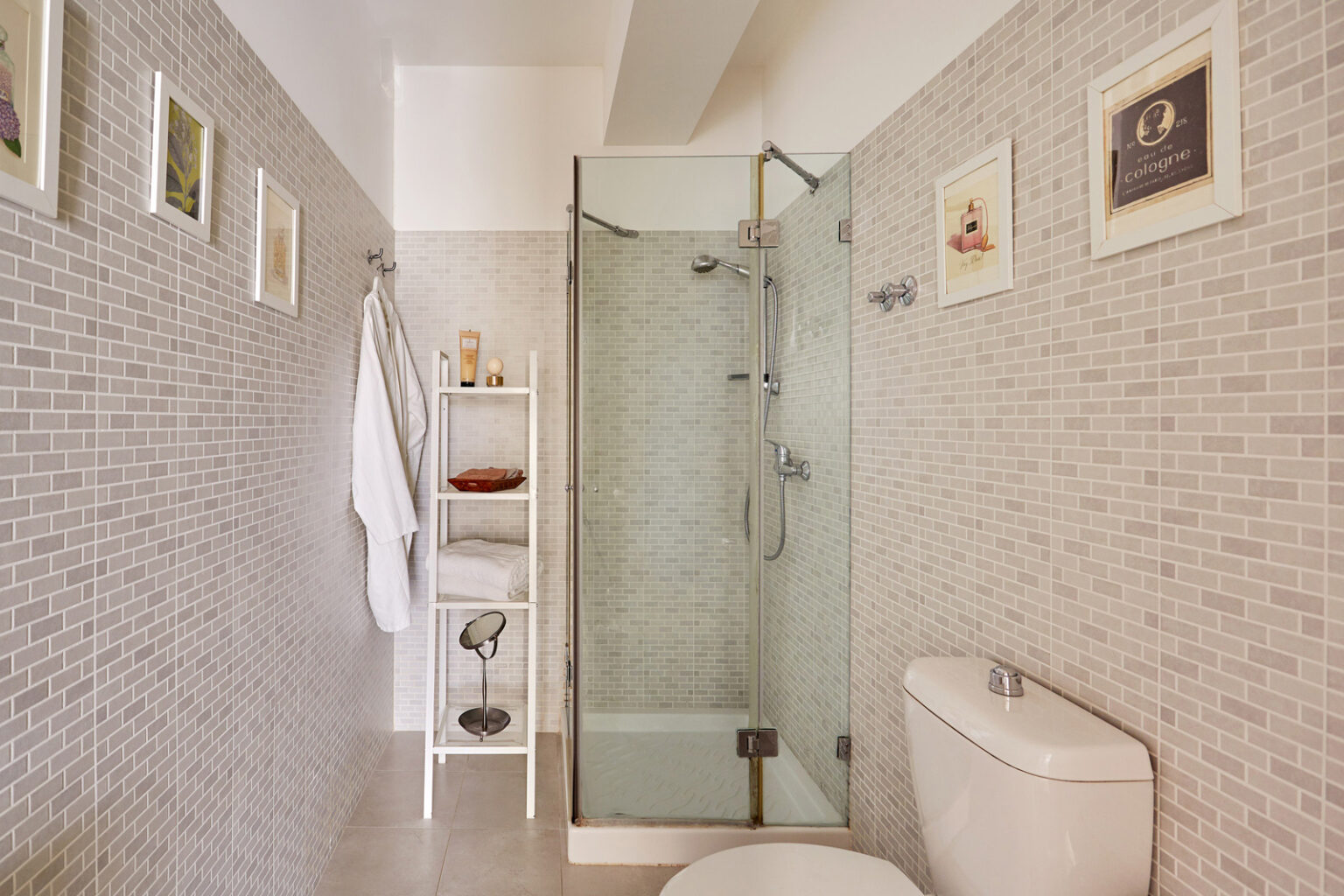 Modern bathroom interior featuring a glass shower, white toilet, and tiled walls. Includes a towel rack, a shelf with toiletries, and framed wall art for a stylish touch. | Only on SopranoVillas