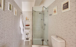 Modern bathroom interior featuring a glass shower, white toilet, and tiled walls. Includes a towel rack, a shelf with toiletries, and framed wall art for a stylish touch.