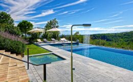 Luxury outdoor pool with a modern shower, sun loungers, and lush greenery under a bright blue sky. Ideal for relaxation and leisure.