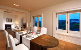 Modern dining room with a wooden table set for a meal, featuring elegant white chairs, candlelight, and large windows showcasing a scenic view. The warm interior design includes soft lighting and a cozy fireplace, creating an inviting atmosphere for dining.