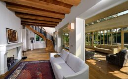 Modern living room with a white sofa, wooden beams, and a staircase, featuring large windows that open to a sunlit area with additional seating and greenery. Ideal for showcasing contemporary interior design and open-concept living spaces.