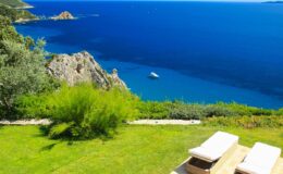 Scenic coastal view featuring lush green grass, two lounge chairs, and a deep blue sea with rocky cliffs and a small boat in the distance. Ideal for relaxation and vacation imagery.