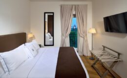Modern hotel room featuring a king-size bed with white linens, a flat-screen TV mounted on the wall, a full-length mirror, and a cozy seating area near a window with curtains. A stylish floor lamp and a luggage rack add to the room's elegant decor.